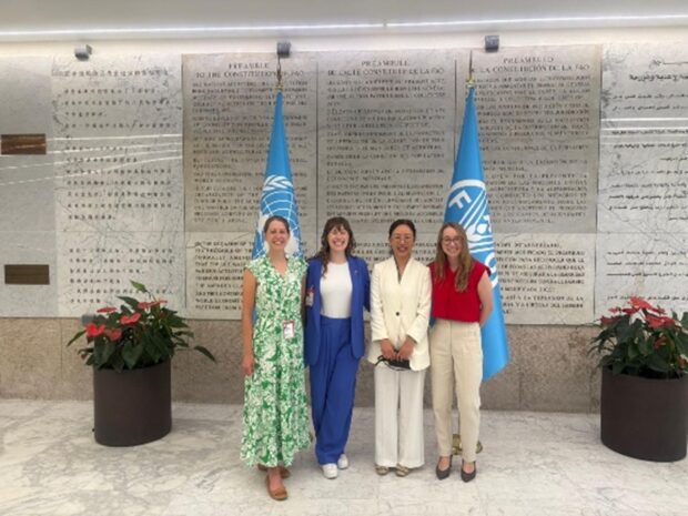 Four women are standing in front of a large wall, with two blue FAO flags and plant pots on either side of them. They are smiling at the camera. 
